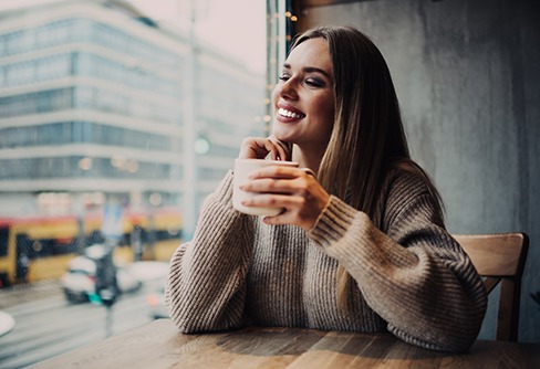 Woman seated at table window holding coffee cup smiling looking at cars passing by